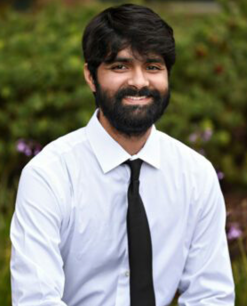 Smiling man with dark hair and beard wearing a white dress shirt and black tie, outdoors in a garden setting.
